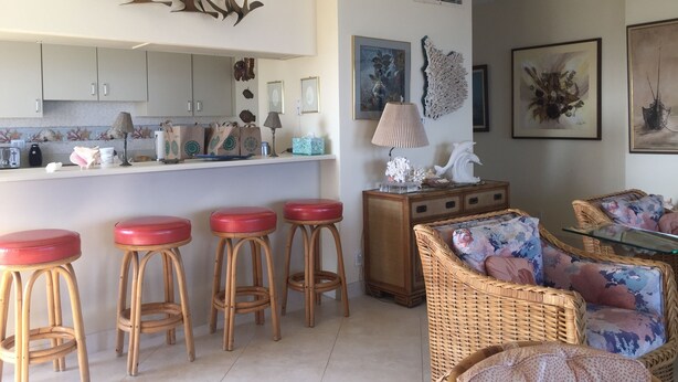 A living room with a kitchen area, wicker furniture, and decorative wall art. -   Three bed room, beach- front  end unit with magnificent views, on the11 floor. (Galveston)