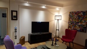 Una sala moderna con televisor de pantalla plana, una silla roja, una silla morada, una lámpara de pie y un cuadro abstracto de colores en la pared.