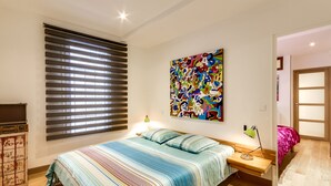 Un dormitorio con cama, un cuadro abstracto colorido, una ventana con persianas y un banco de madera.