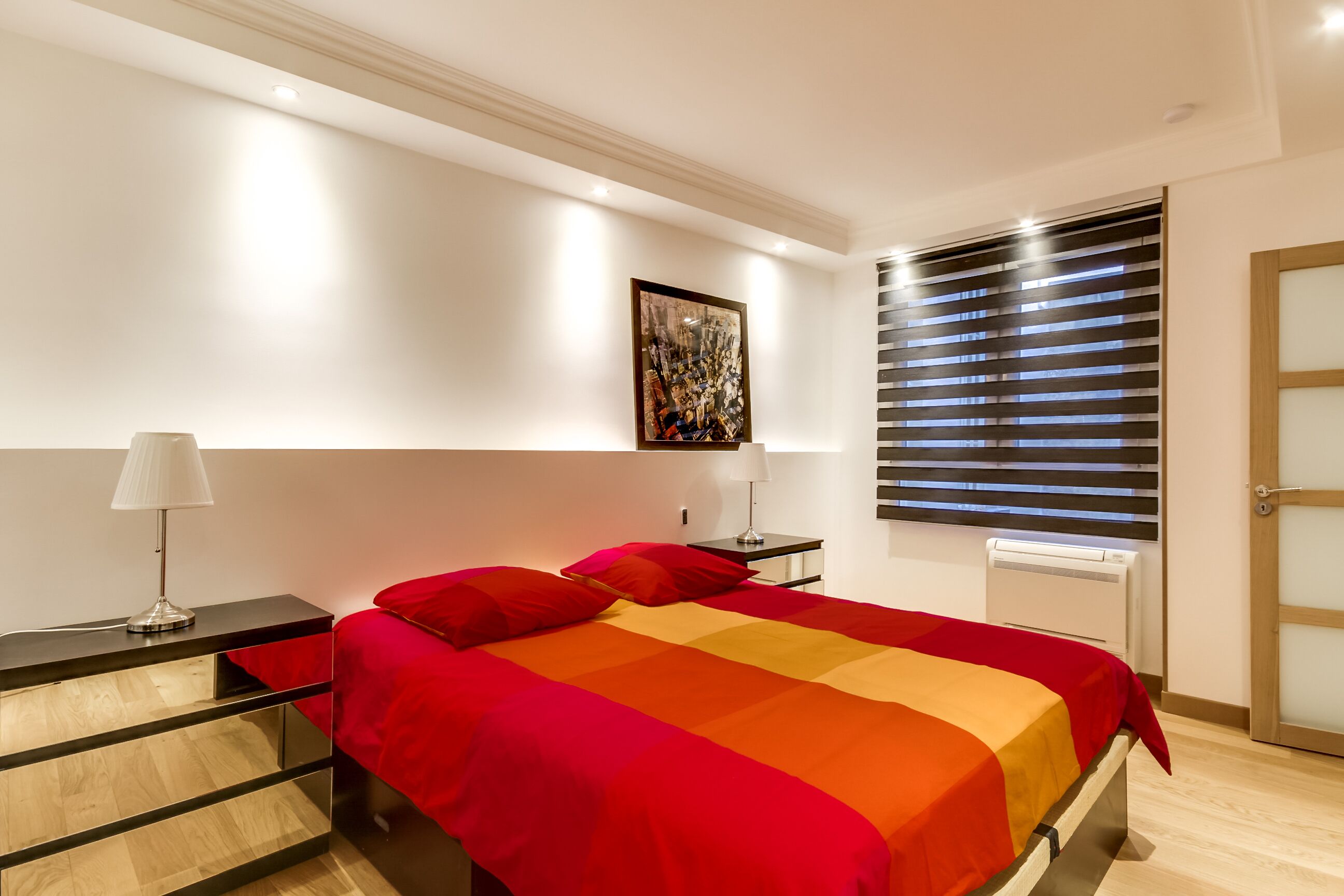 Une chambre moderne avec un lit, des tables de chevet, une œuvre d’art accrochée au mur et une fenêtre avec des stores.