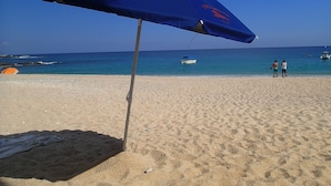 Een strand met een blauwe parasol, een boot en twee personen die op het zand staan.