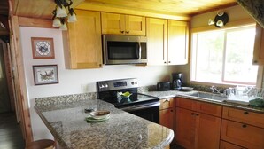 Una cocina con armarios de madera, encimeras de granito, un microondas, estufa y un lavabo. Hay dos taburetes y un cubo de basura.