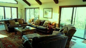 A living room with leather sofas, a coffee table, and a large rug. There are multiple windows providing natural light and a view of the outdoors.