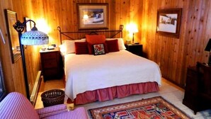 A wooden-paneled bedroom with a bed, two bedside tables, a floor lamp, a framed picture on the wall, and a patterned rug.