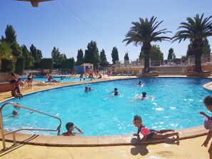 A swimming pool with people swimming and playing, surrounded by palm trees and a clear sky.
