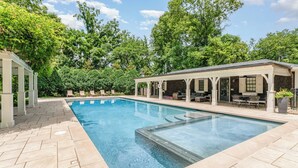 Una piscina rectangular con un borde de baldosas, un porche cubierto con mobiliario exterior y abundante vegetación al fondo.