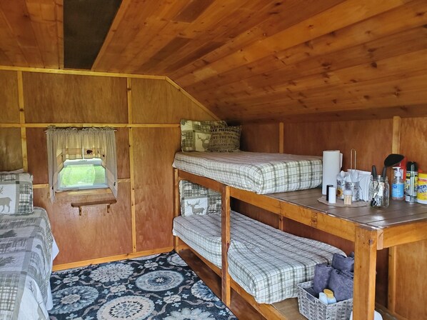 A wooden cabin room with bunk beds, a window with curtains, a patterned rug, and various items on the table.
