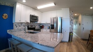 A kitchen with white cabinets, a stainless steel refrigerator, a microwave, and a sink. There are three white bar stools and a wooden dining table. - “BEACHFRONT ELM” IS CALLING
~Oceanfront~Private Beach Access~Pool~ (Carolina Beach)