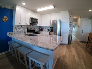 A kitchen with white cabinets, a stainless steel refrigerator, a microwave, and a sink. There are three white bar stools and a wooden dining table.