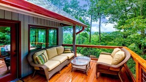 An outdoor deck with wicker furniture, a glass table, and a view of a forested area. - Sweet Refrain in Pisgah Forest with VIEW (PIsgah Forest)