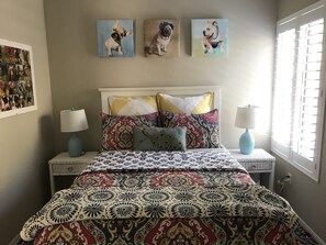 A bedroom with a patterned bedspread, decorative pillows, a framed collage of dogs, and two bedside lamps.