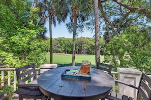 Une table ronde avec une carafe, un plateau et un livre, entourée de chaises, installée à l’extérieur, avec des arbres et un terrain de golf en arrière-plan.