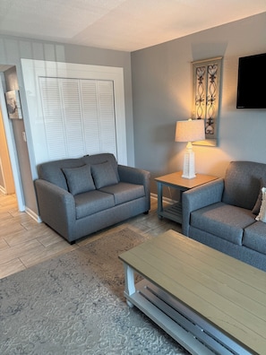 A living room with a grey sofa, a matching armchair, a small table with a lamp, and a wooden coffee table.