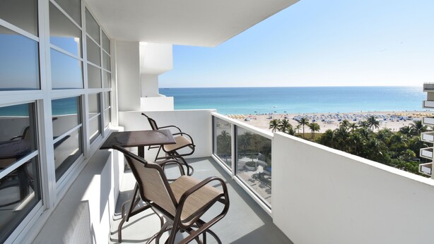 A balcony with a table and chairs, overlooking a beach with palm trees and a clear sky. - Between Lowe's and Ritz Carlton Hotels Beachfront on Lincoln (Miami)