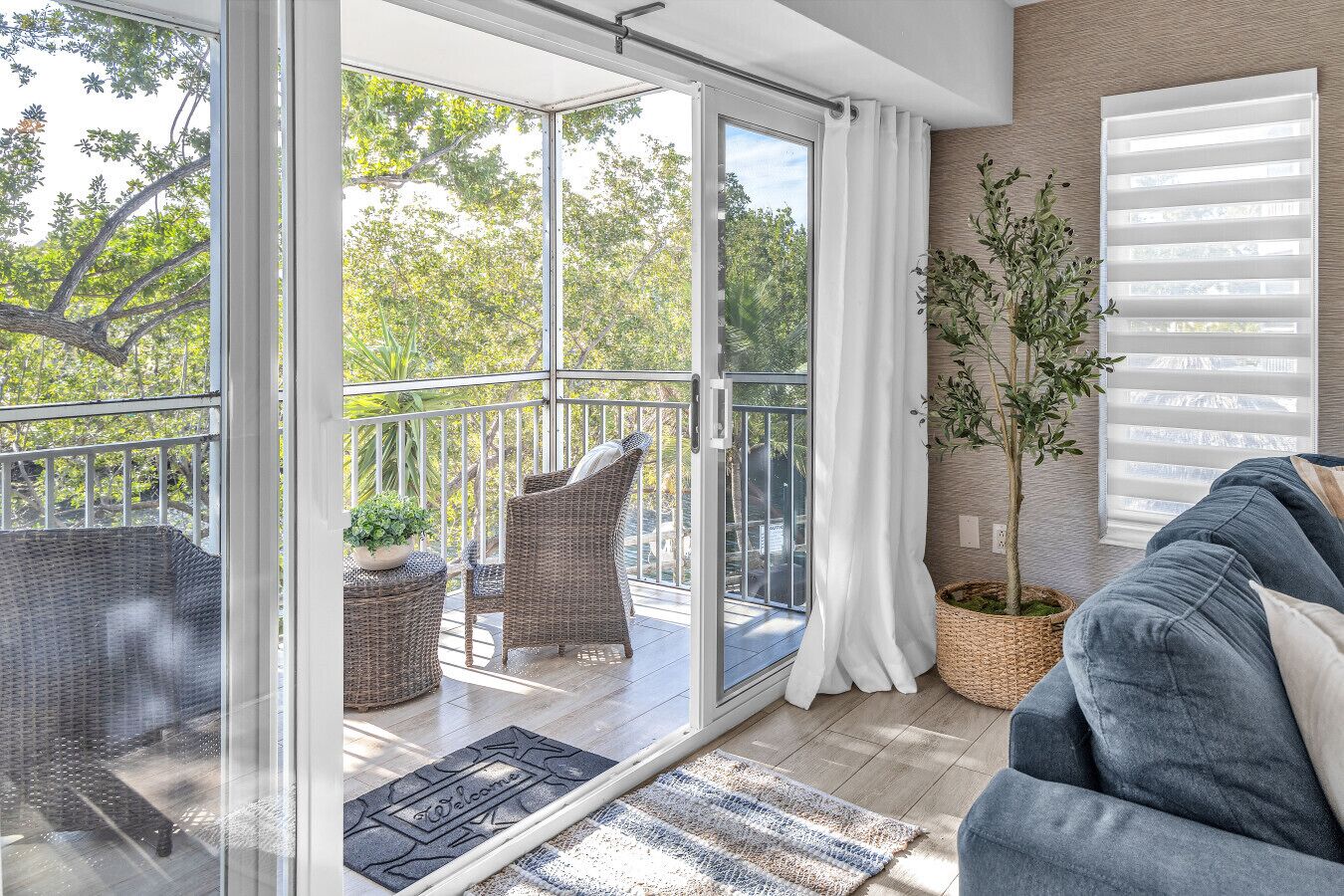 A room with a sliding glass door leading to a balcony, wicker furniture, a potted plant, and a blue sofa.
