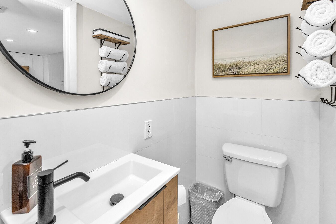 A bathroom with a white sink, a toilet, a trash bin, a mirror, and a towel rack with towels.