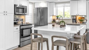 A modern kitchen with white cabinetry, stainless steel appliances, a central island with bar stools, and a large window providing natural light. - Key Largo Kawama Villa L1, 3/3 (Key Largo)