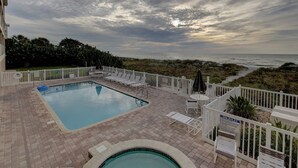 A pool area with a view of the ocean, surrounded by a white fence and lounge chairs.