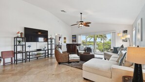 A spacious living room with a large TV, a ceiling fan, and a view of palm trees through large windows.