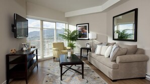 A living room with a beige sofa, a coffee table, a TV mounted on the wall, a potted plant, and a window with blinds. - Lake View! Resort Style living. Short term rentals are allowed  at this complex. (Westbank, Kelowna)