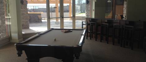 A pool table in a room with large windows, a bar area with stools, and a stone wall.