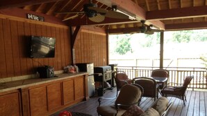 A covered outdoor seating area with wooden furniture, a television mounted on the wall, and a grill.