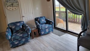A room with two blue armchairs, a wall clock, a door, and a view of a wooden deck through a sliding glass door.