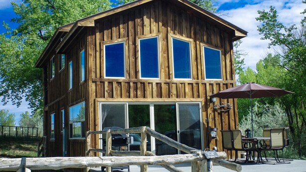 Una cabaña de madera con ventanas amplias, una terraza y mobiliario exterior.