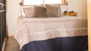 Un dormitorio con una cama, una ventana con persiana, una mesita de noche con lámpara y un estante en la pared.