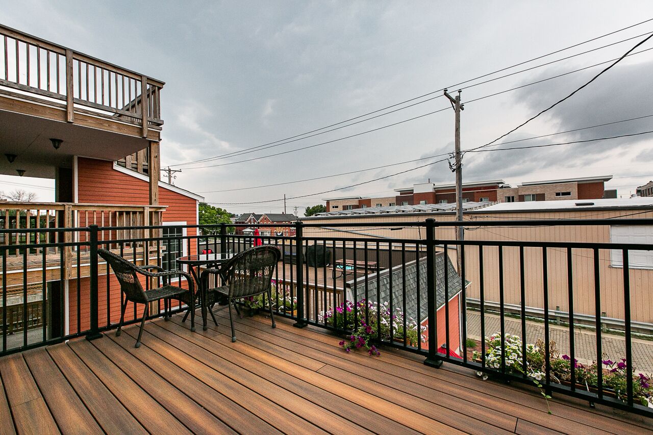 Une terrasse en bois avec deux chaises et une table, donnant sur un quartier résidentiel avec des maisons et un immeuble à la façade rouge.