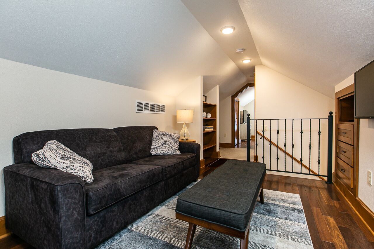 Un salon avec un canapé foncé, un banc et une lampe. On y trouve un escalier avec une rampe menant à un niveau supérieur.