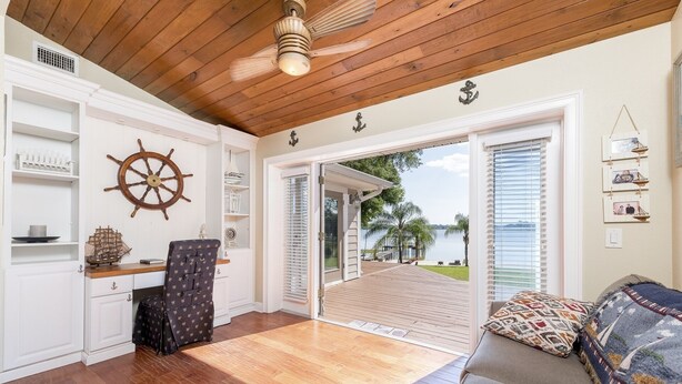 A room with a wooden ceiling, a ship's wheel, a desk with a chair, a sofa with patterned cushions, and a view of a body of water through a door.
