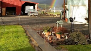 Ländliche Szenerie mit einem roten Bauernhaus, einem weißen Lastwagen und einer Garage. Im Hintergrund ist ein Regenbogen sichtbar.
