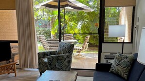 A living room with a sofa, a coffee table, and a TV. There is a large window with a view of greenery outside. - UPDATED MAUI VACATION RENTAL (Kihei)