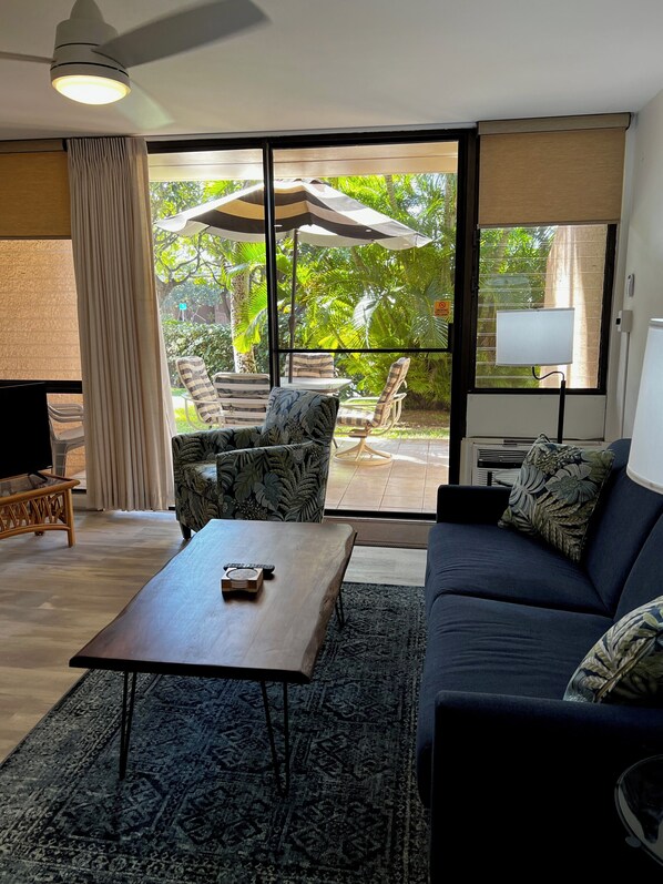 A living room with a sofa, a coffee table, and a TV. There is a large window with a view of greenery outside.