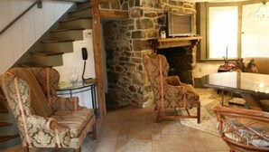 A cozy living room with a stone fireplace, a television mounted above it, and a dining area with a table and chairs. - Cottage on Historic Gentleman's Farm (Chester Springs)