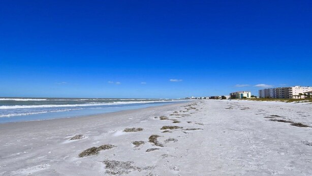 A beach with footprints leading to the ocean, buildings in the background, and a clear blue sky. - Beautifully Updated Madeira Bay Resort Unit 401 - Steps from Sand and Fun! (Madeira Beach)