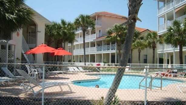 A pool area with lounge chairs and umbrellas, surrounded by a fence, with a building in the background.