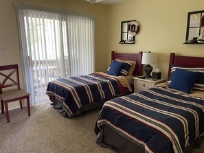 Habitación de hotel con dos camas, una silla, una mesita de noche con lámpara y una ventana con persianas.