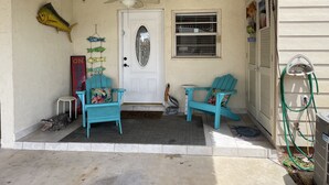 A porch with two turquoise chairs, a ceiling fan, string lights, a fish decoration, a door with a frosted glass panel, and a window with a metal grill.