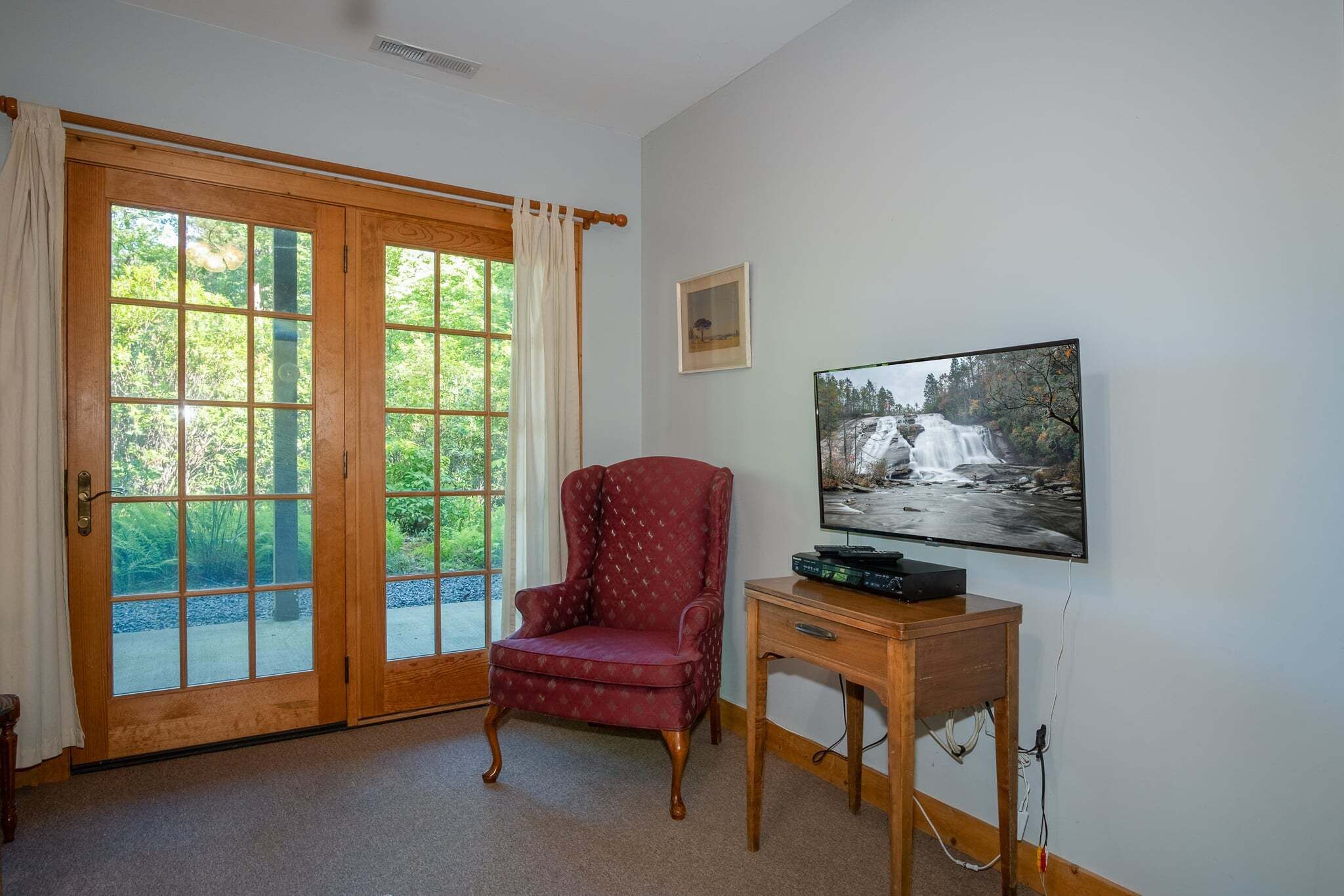 Una habitación con una silla de madera, un televisor sobre un soporte de madera y un amplio ventanal con vistas a la vegetación.