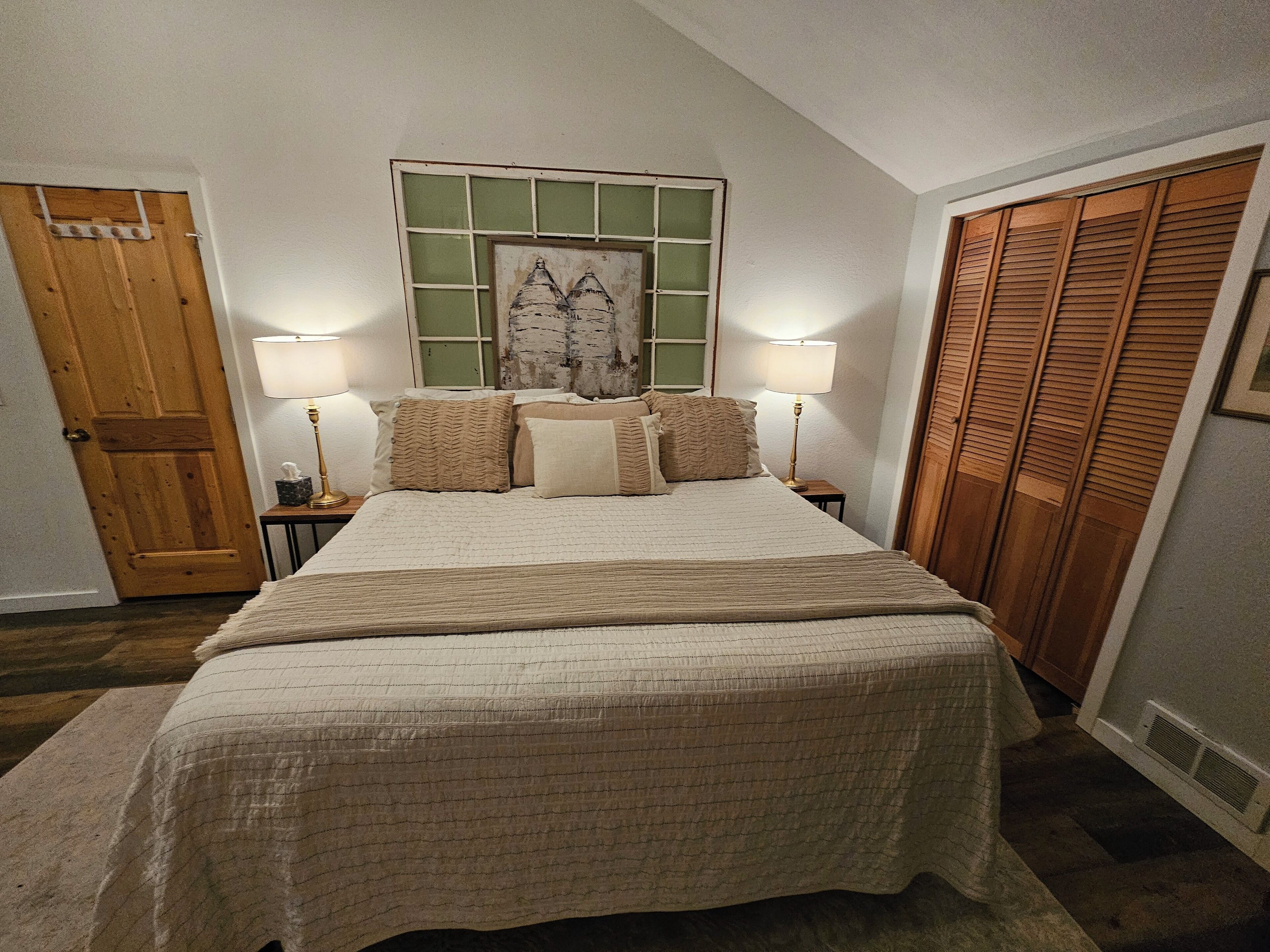 Un dormitorio con una cama grande, dos lámparas de noche, una obra de arte con marco verde sobre la cama y un armario de madera.