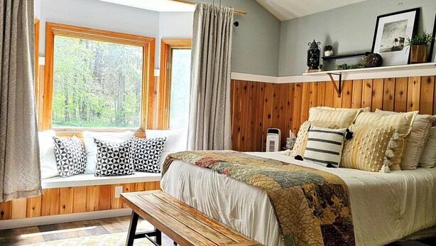 Een slaapkamer met een bed, een bankje, een plafondventilator, een raam met gordijnen en een houten wand.