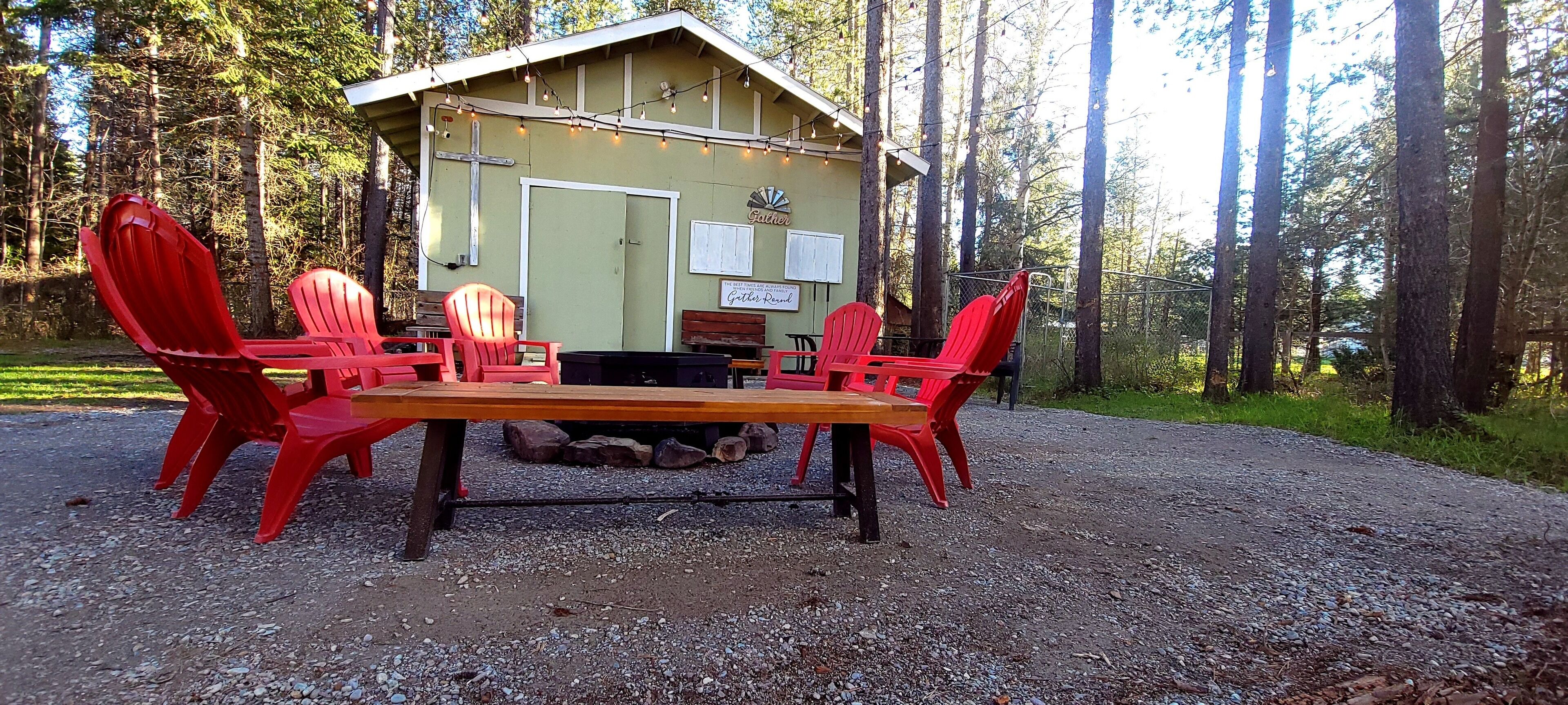 A rustic outdoor seating area with a wooden table and red Adirondack chairs, surrounded by a gravel path and a forested backdrop.
