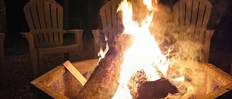 A fire pit with two Adirondack chairs around it, surrounded by trees and string lights.