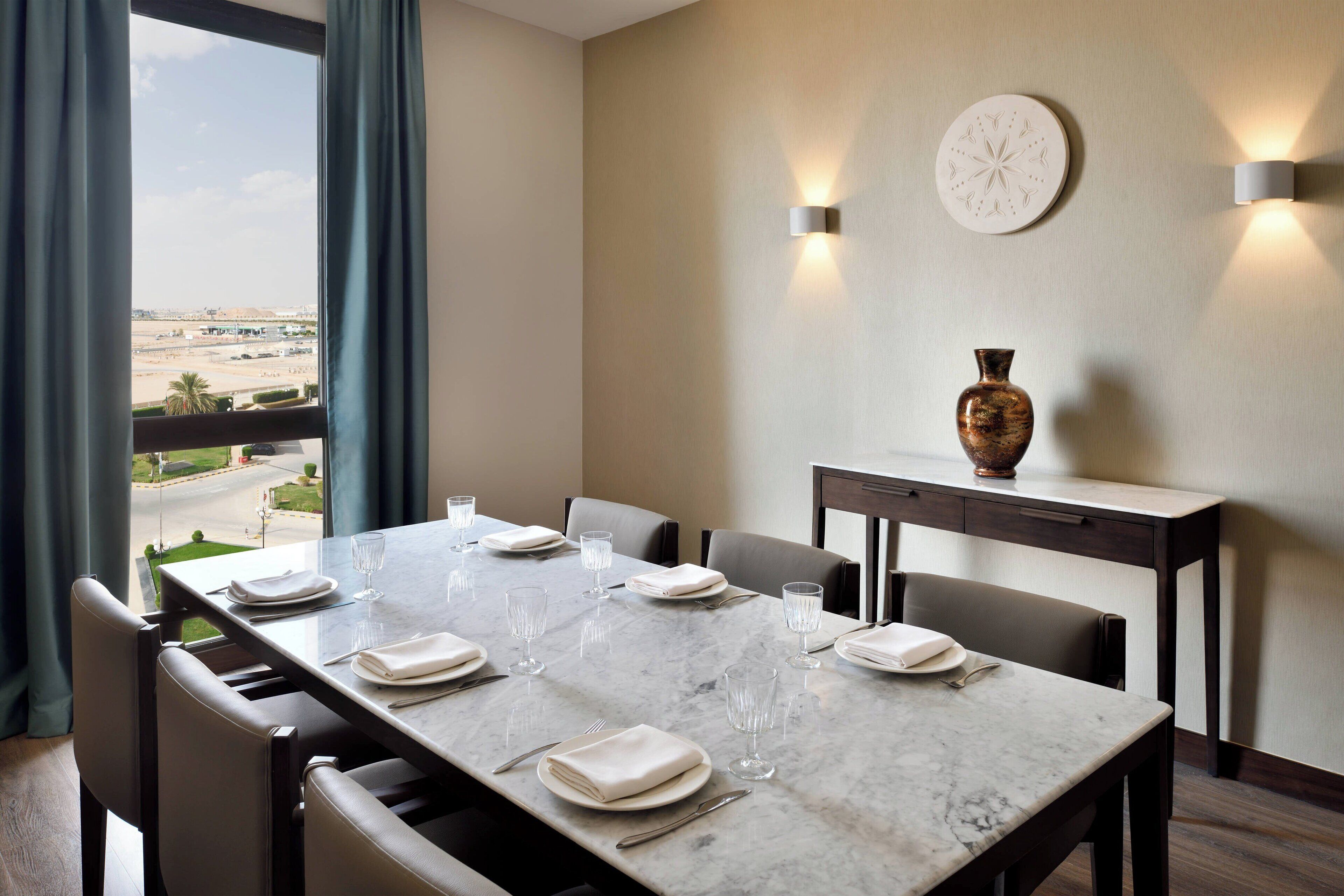 A dining area with a marble table set for six, a vase on a side table, and a view of a desert landscape through large windows.