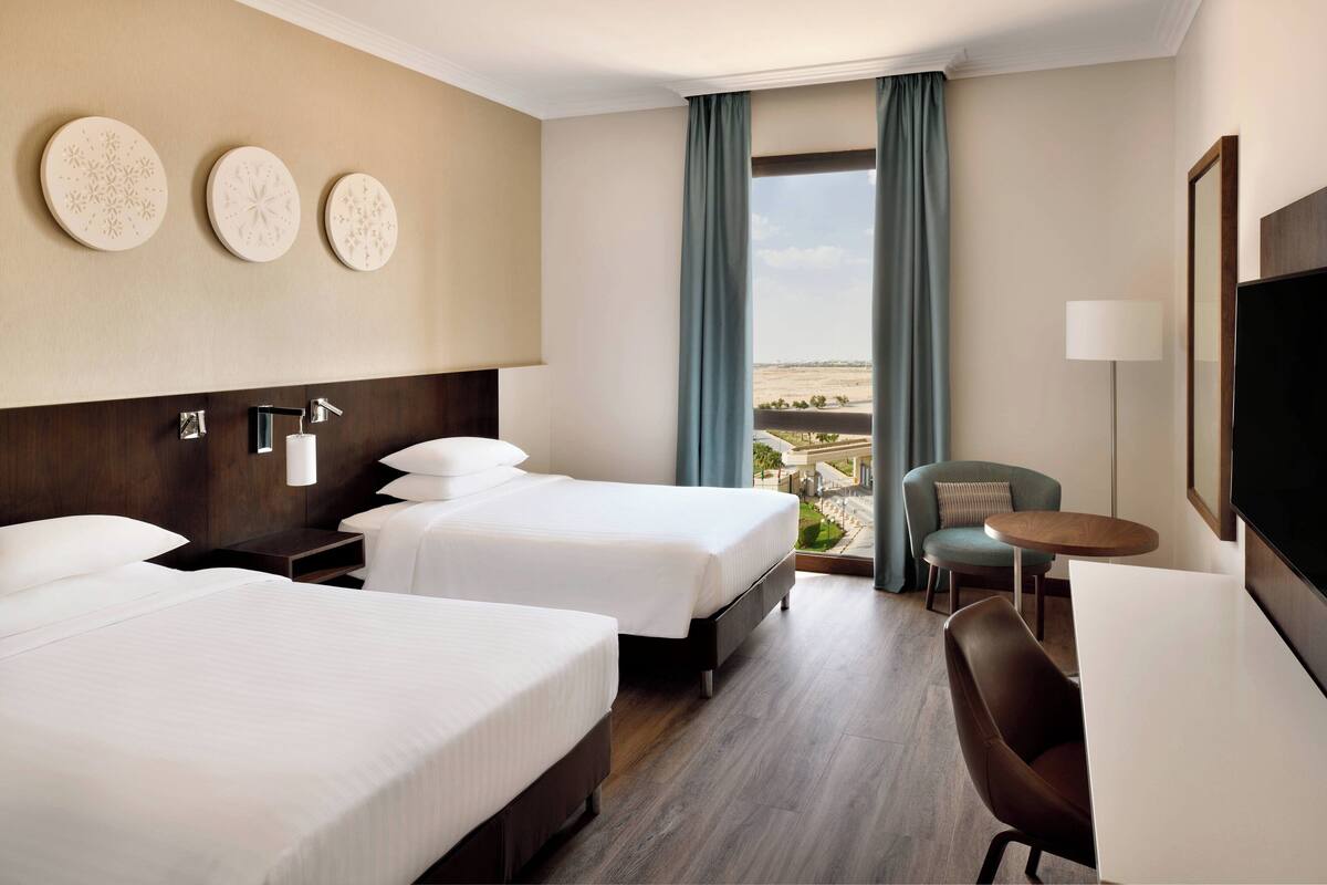 Ein Hotelzimmer mit zwei Betten, einem Schreibtisch, einem Stuhl und Blick auf die Landschaft.