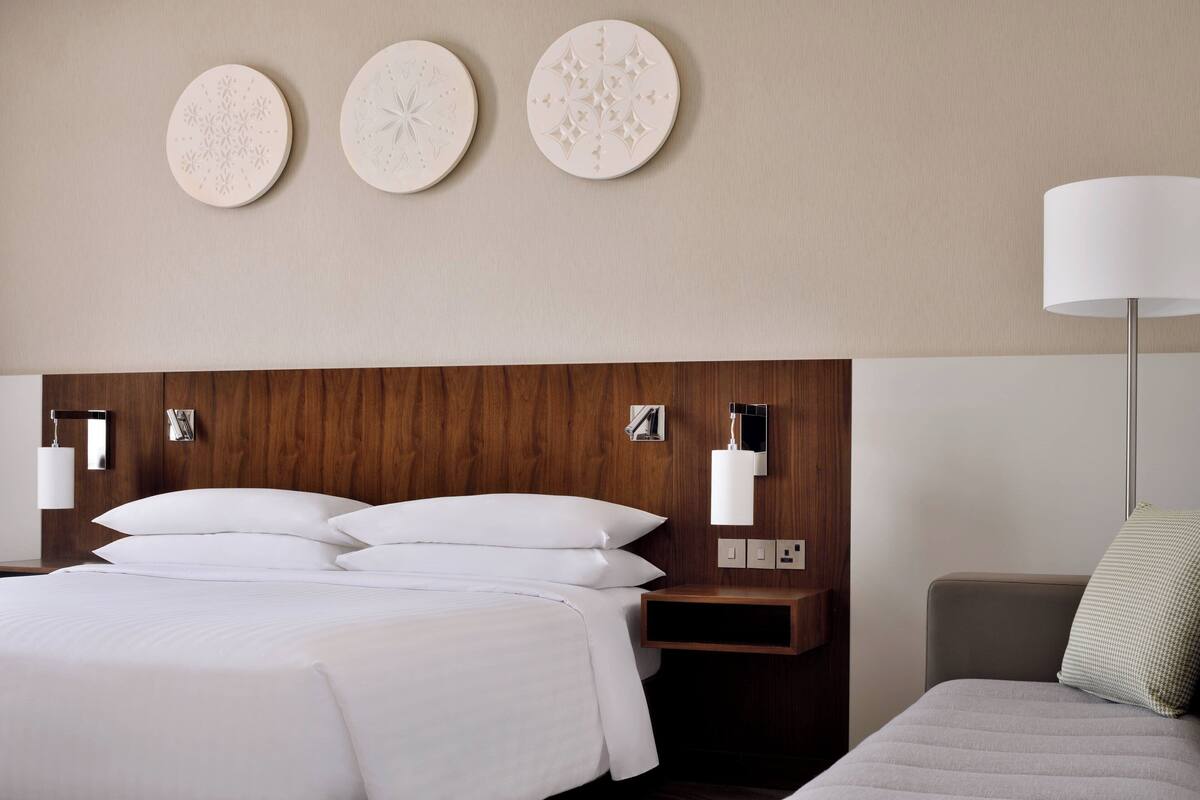 Ein Hotelzimmer mit einem hölzernen Kopfteil, einem Bett mit weißer Bettwäsche, einem Nachttisch mit Lampe und drei an der Wand befestigten Dekorationsplatten.