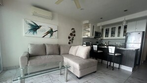 Una sala de estar moderna con una mesa de centro de vidrio, un sofá seccional color beige y una cocina con refrigerador, microondas y encimeras oscuras.