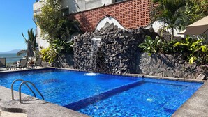 Una piscina con agua azul cristalina, rodeada de un muro de piedra y plantas tropicales.